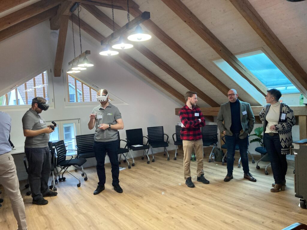 Eine Gruppe von fünf Personen zu sehen bei der Gruppenarbeit bei der Silversgroup Zwei Personen verwenden VR-Headsets, während drei andere in der Nähe stehen und sich unterhalten. Im Hintergrund stehen leere Stühle an der Wand.
