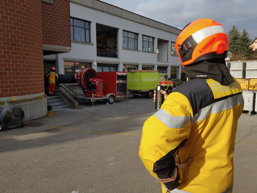 Ein Feuerwehrmann der Ortsfeuerwehr in gelber Jacke und orangefarbenem Helm beobachtet ein Team bei der Arbeit in der Nähe eines Gebäudes. Geräte wie Lüftungsgeräte und ein Anhänger sind zu sehen. Das Gebäude hat große Fenster und die Szene spielt im Freien.